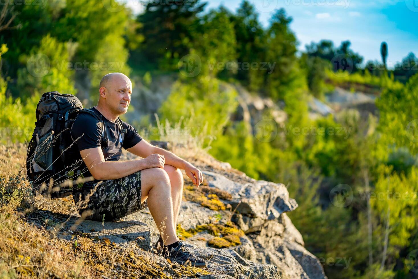 Traveler looks at the landscape around himself at the top of the cliff in the summer in warm weather. A man sits on squat near a cliff in a sunny day photo