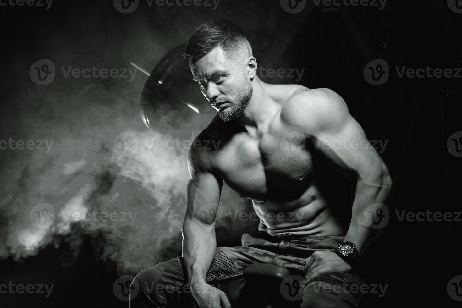 Bodybuilder posing on black background. Sitting on bench after training. Muscular man in gym. Black and white photo