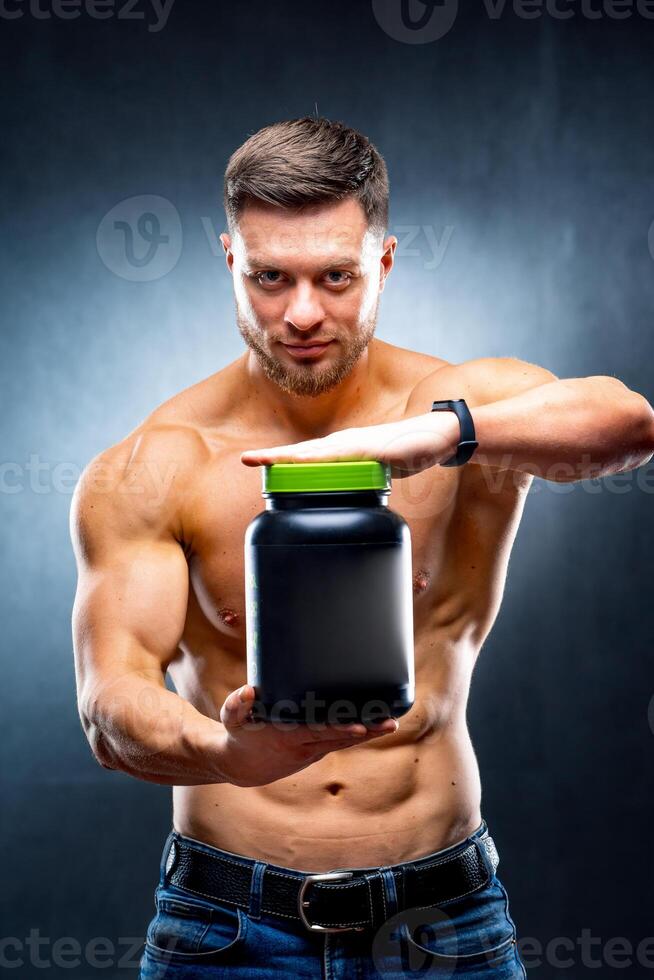 Male athlete bodybuilder holds a jar of sports nutrition in his hands showing to the camera poses on blue background. photo