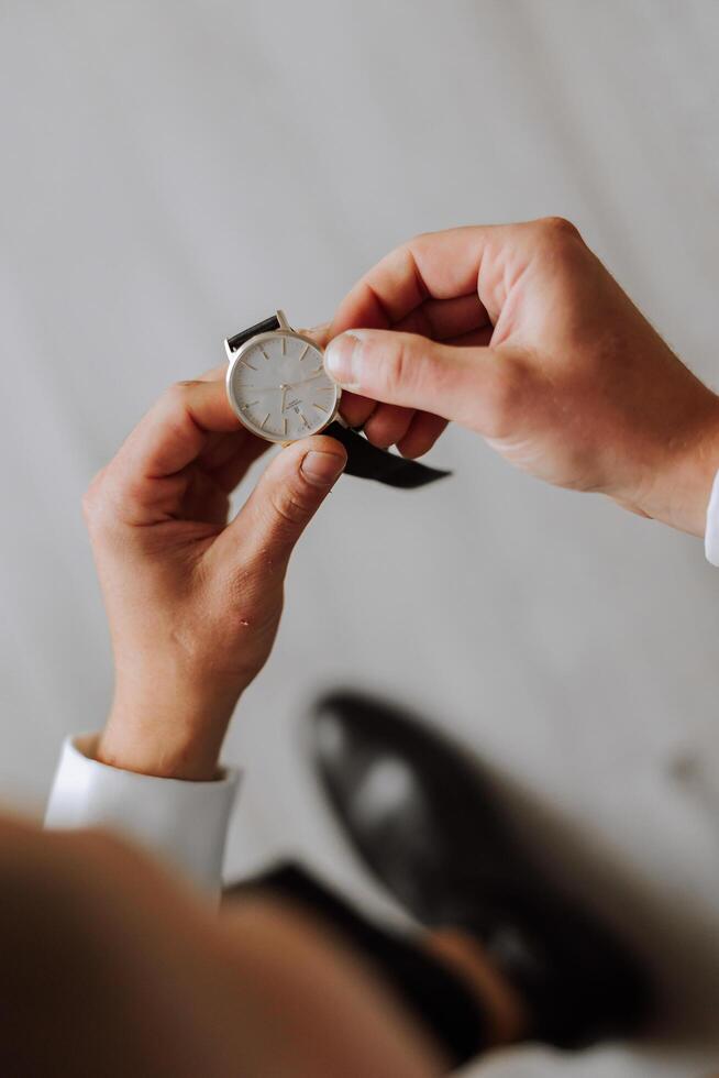un de cerca de un recortado marco de un hombre pone en un reloj con un cuero cinturón, es vestido en un elegante traje, un blanco camisa, usa un oro anillo. foto