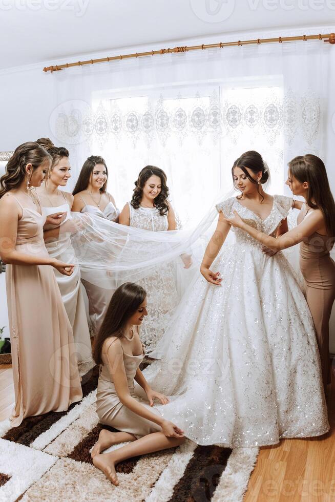 Bridesmaids rejoice in the morning, helping to fasten the buttons on the wedding dress and prepare for the wedding ceremony. They take pictures, smile, help the bride with her shoes. photo