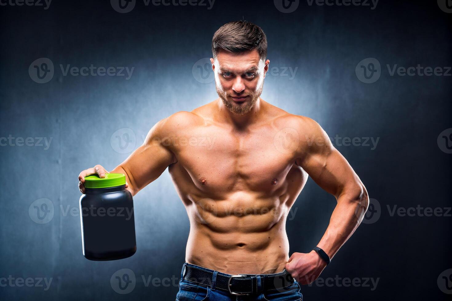 Male athlete bodybuilder with a naked torso holds a jar of sports nutrition in his hands and poses on a blue background. photo