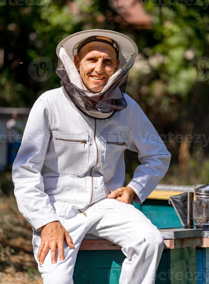 hermoso adulto hombre apicultor en blanco protector traje a colmenar. verano jardín con urticaria borroso antecedentes. de cerca. foto