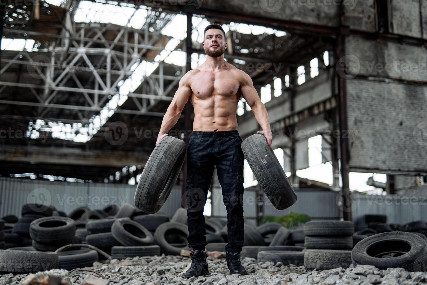 Strong young man posing at the abandoned place with tires at the background. Big muscles and perfect abs. Many tires on the background. Strongman after workout photo