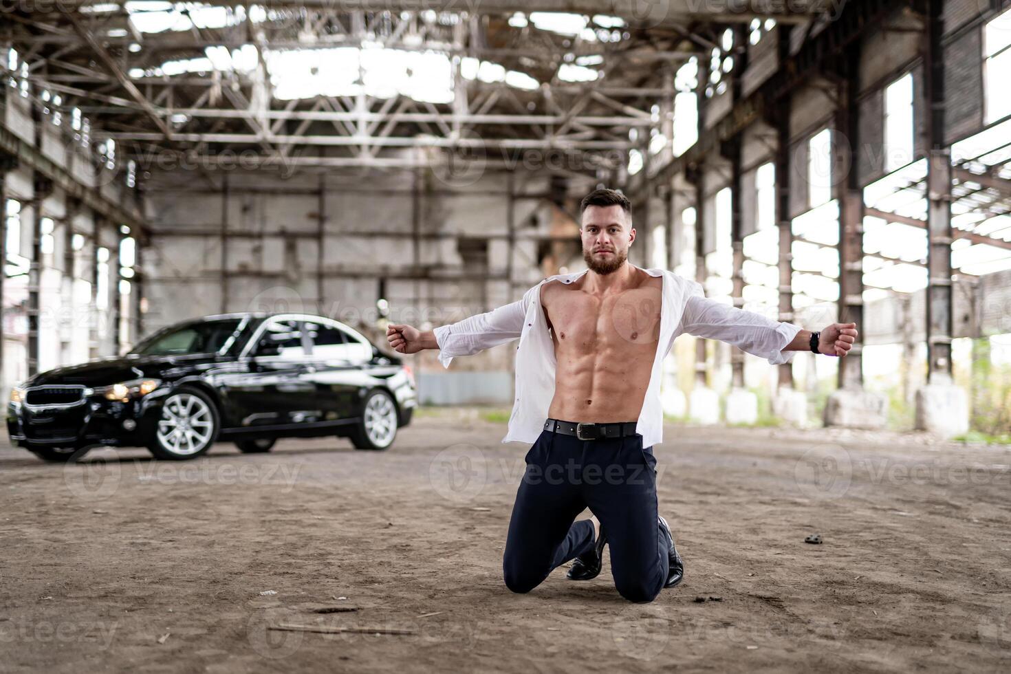 Rich man in unbuttoned white shirt standing at the knees and posing with the luxury car at the background. Black auto at abandoned warehouse photo