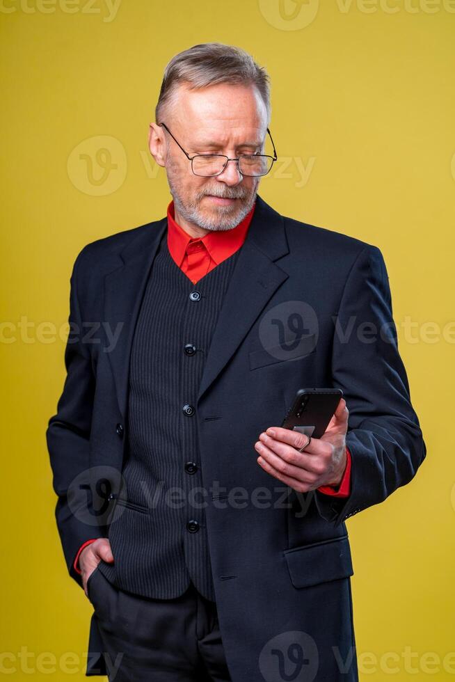 Portrait of senior businessman holding in hands phone while standing at studio over yellow background. Business people concept. photo