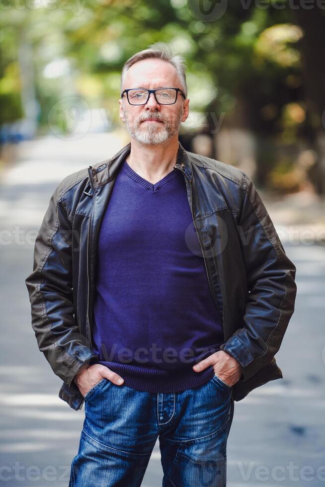 hermoso medio años hombre con gris pelo en un al aire libre ajuste. cuero chaqueta. azul suéter. los anteojos. barbado confidente empresario. borroso antecedentes. manos en bolsillos foto