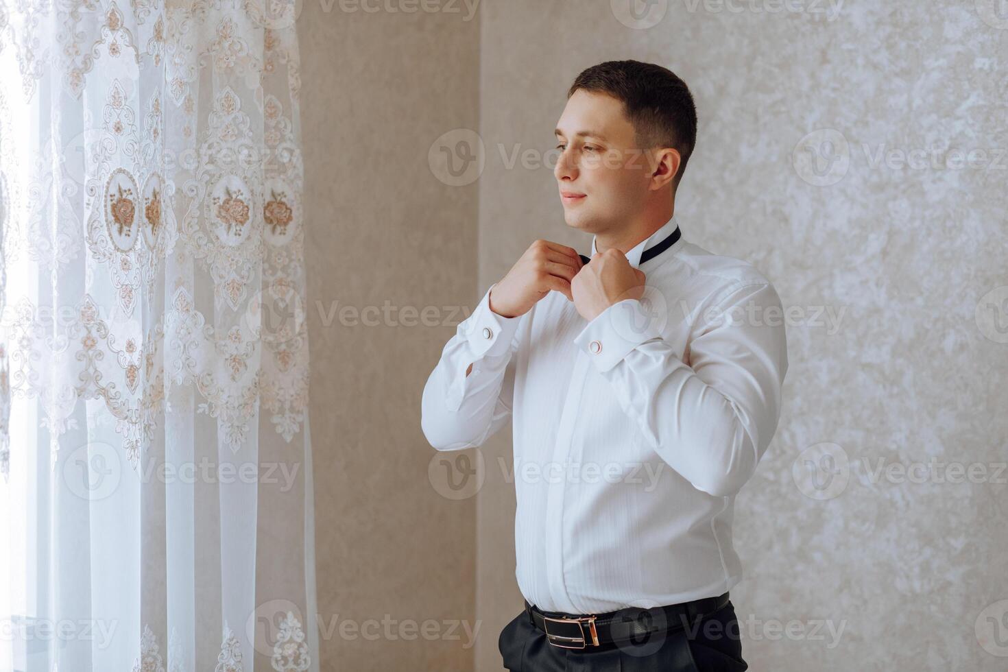 a young man in a white shirt stands by the window in the room and puts on a tie. The groom gets dressed in the morning and prepares for the wedding. photo