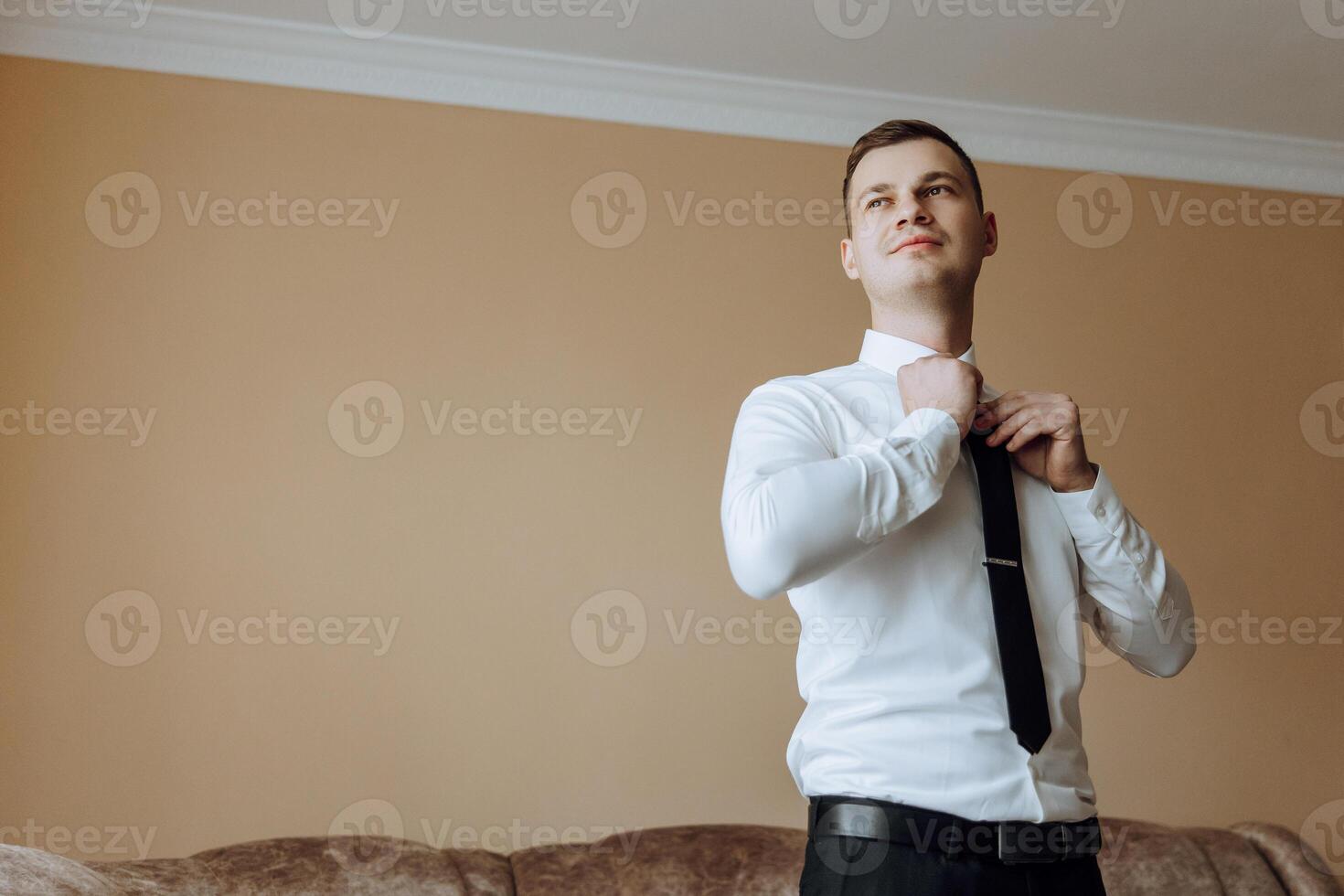a young man in a white shirt stands by the window in the room and puts on a tie. The groom gets dressed in the morning and prepares for the wedding. photo