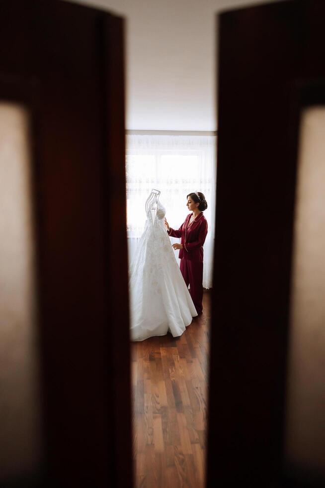 morning portrait of a luxurious and beautiful bride in a hotel in a luxurious room in red pajamas standing next to her wedding dress. photo