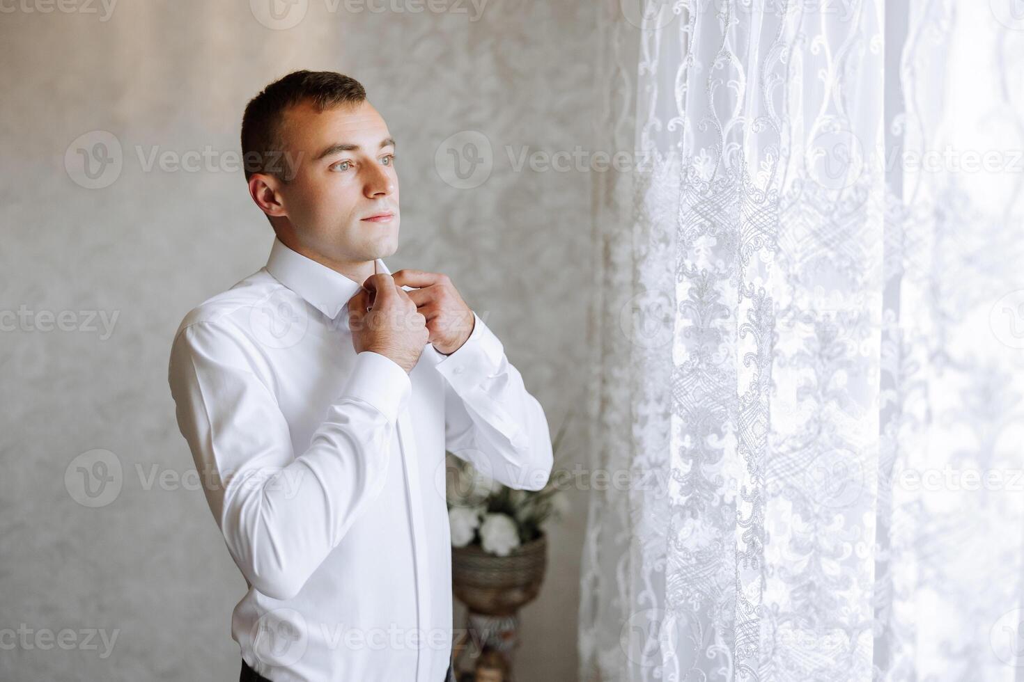 A man in a white shirt stands by the window in the room and fastens the buttons on his collar and sleeves. Watch on hand. Stylish business portrait of a man, close-up photo. The groom is preparing. photo