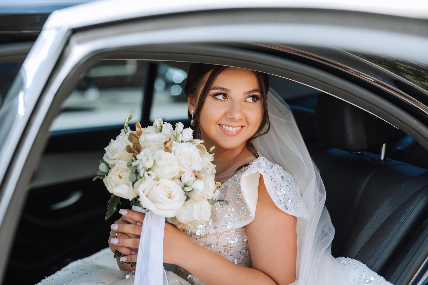 el novia es sentado en un negro coche con un ramo de flores de flores en su manos y mira fuera de el coche. retrato de un bastante tímido novia en un coche. novia, sonrisa, emociones foto