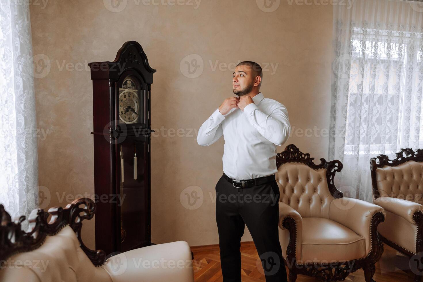 A man in a white shirt stands by the window in the room and fastens the buttons on his collar and sleeves. Watch on hand. Stylish business portrait of a man, close-up photo. The groom is preparing. photo