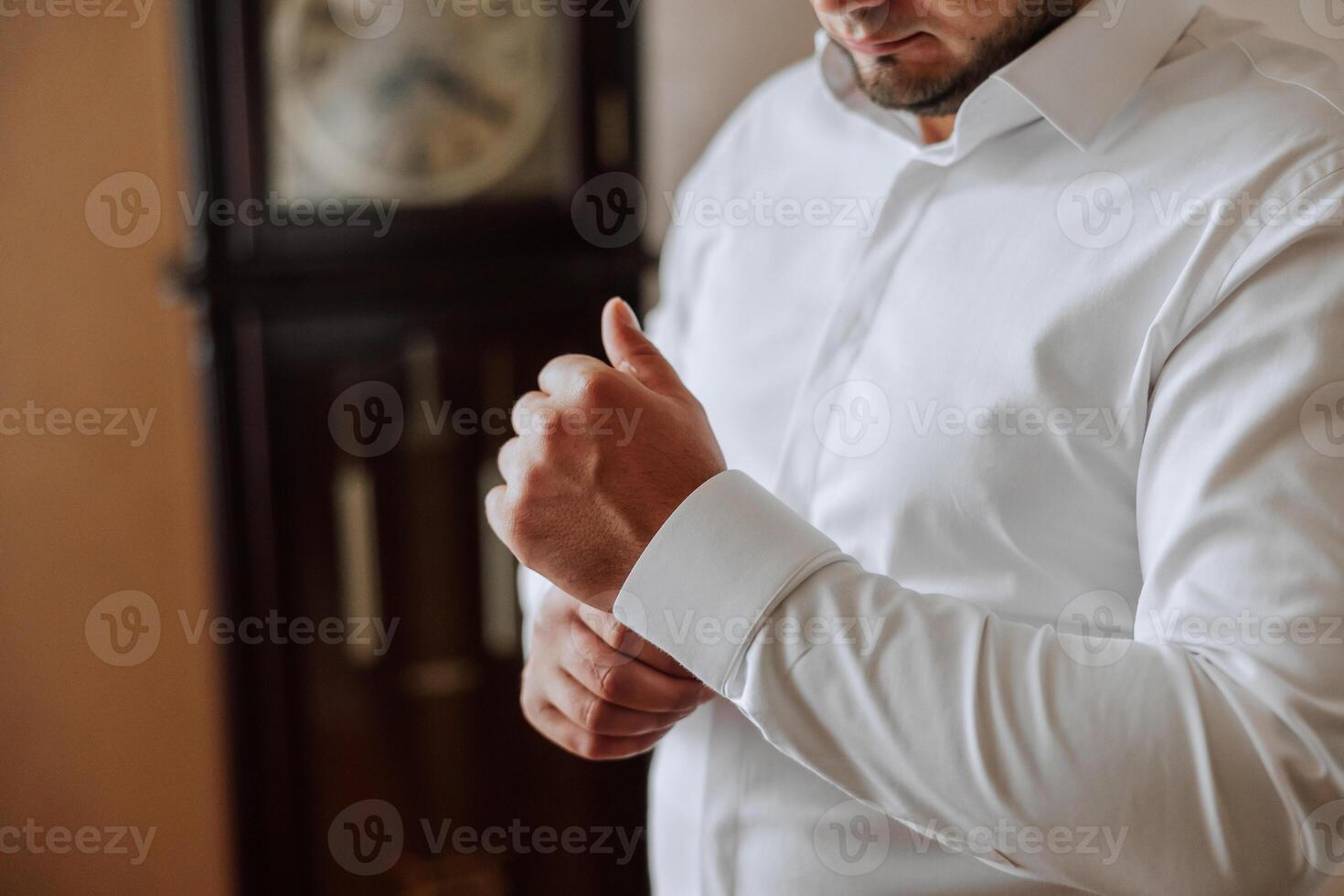 A man in a white shirt stands by the window in the room and fastens the buttons on his collar and sleeves. Watch on hand. Stylish business portrait of a man, close-up photo. The groom is preparing. photo