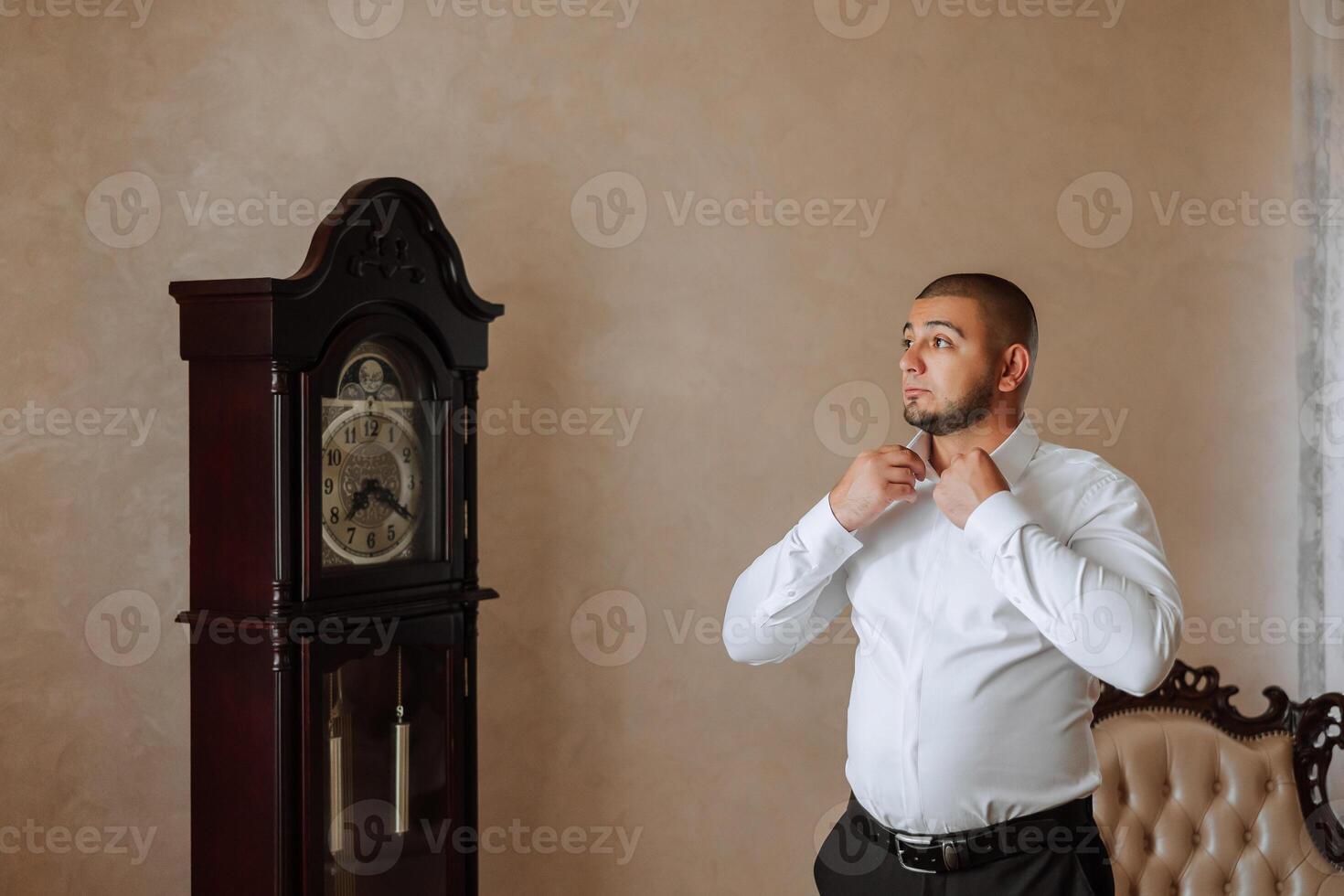 A man in a white shirt stands by the window in the room and fastens the buttons on his collar and sleeves. Watch on hand. Stylish business portrait of a man, close-up photo. The groom is preparing. photo