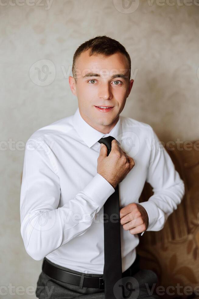 a young man in a white shirt stands by the window in the room and puts on a tie. The groom gets dressed in the morning and prepares for the wedding. photo