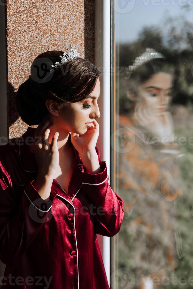 The morning of a luxurious and beautiful bride in a hotel in a luxurious room in red pajamas, standing by the window. photo