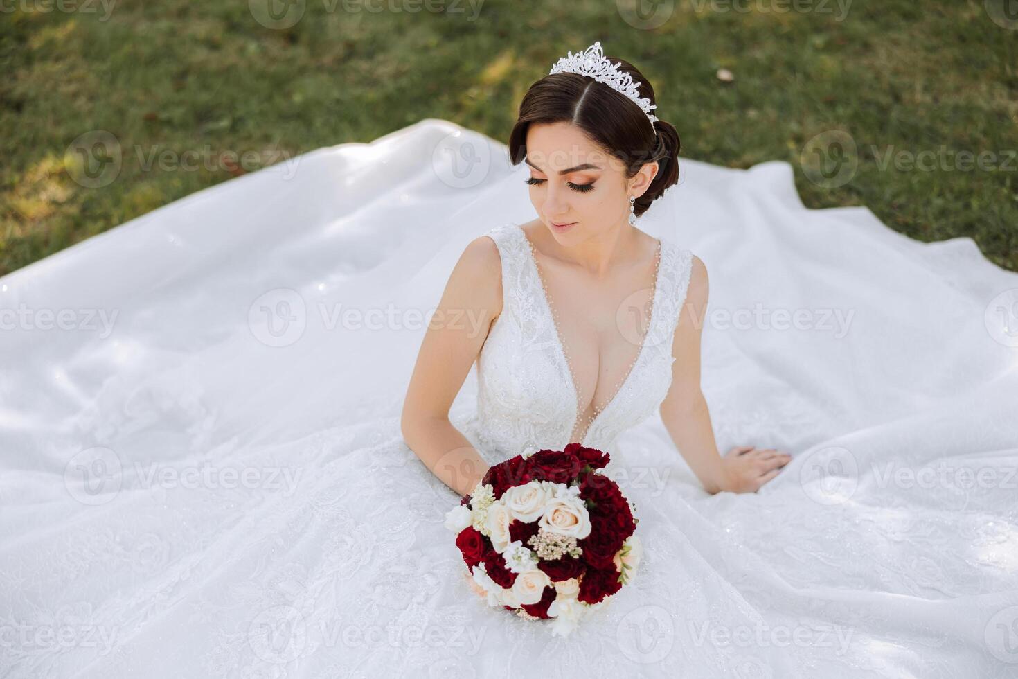A brunette bride with a tiara in her hair, sitting on the ground in a dress unfolded in the shape of a circle, holding a bouquet. On a green background. Sunny day. Wedding ceremony photo