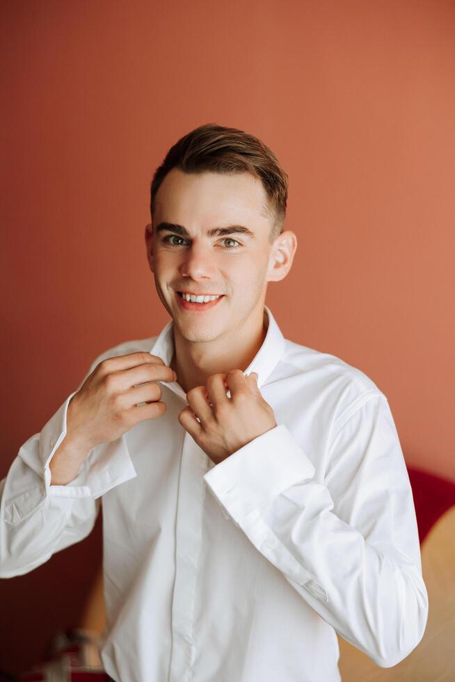 un hombre en un blanco camisa soportes por el ventana en el habitación y sujeta el botones en su collar y mangas reloj en mano. elegante negocio retrato de un hombre, de cerca foto. el novio es preparando. foto