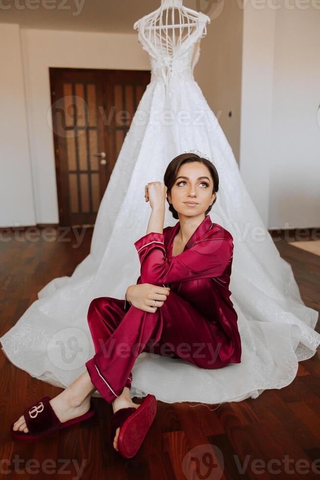 morning portrait of a luxurious and beautiful bride in a hotel in a luxurious room in red pajamas sitting and posing next to her wedding dress. photo