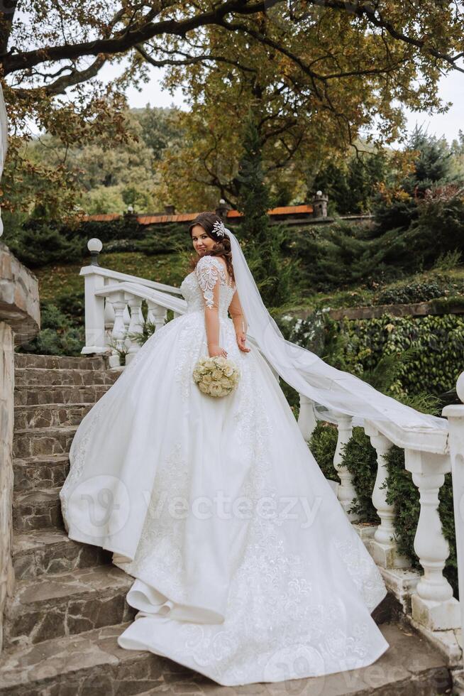 A brunette bride in a white dress with a long train holds the dress and walks down the stone path. Autumn. Wedding photo session in nature. Beautiful hair and makeup. celebration