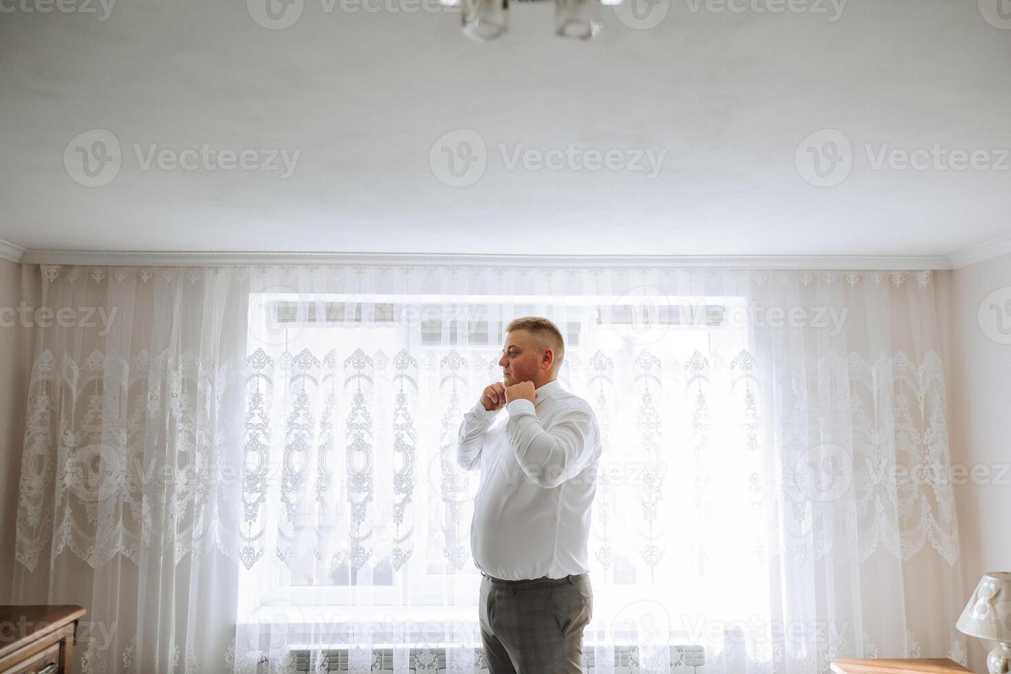 A man in a white shirt stands by the window in the room and fastens the buttons on his collar and sleeves. Watch on hand. Stylish business portrait of a man, close-up photo. The groom is preparing. photo