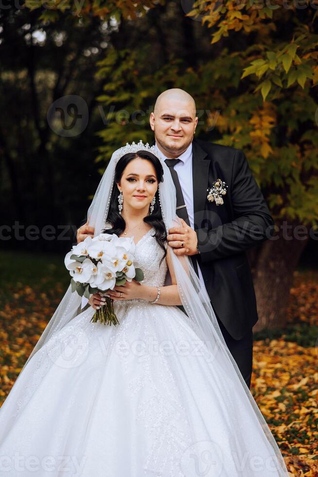 Stylish, young groom and beautiful bride in a long white dress and a long veil with a bouquet in their hands, hugging in the park in the autumn nature. Wedding portrait of newlyweds. photo
