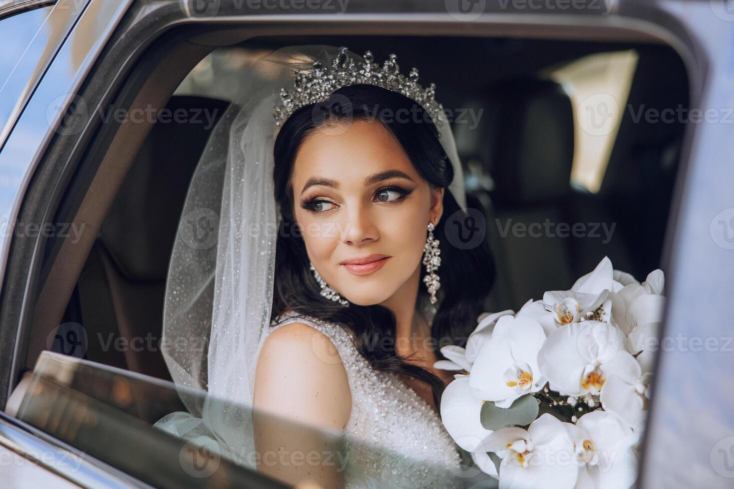 The bride is sitting in a black car with a bouquet of flowers in her hands and looks out of the car. Portrait of a rather shy bride in a car. Bride, smile, emotions. photo