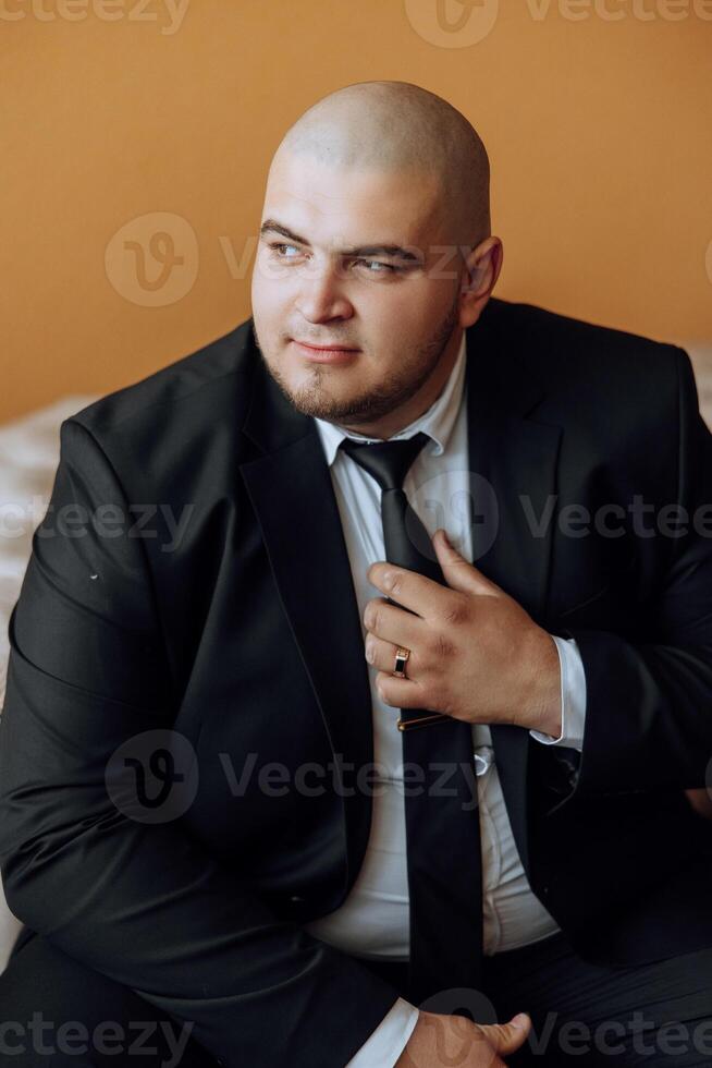 a young man in a white shirt stands by the window in the room and puts on a tie. The groom gets dressed in the morning and prepares for the wedding. photo