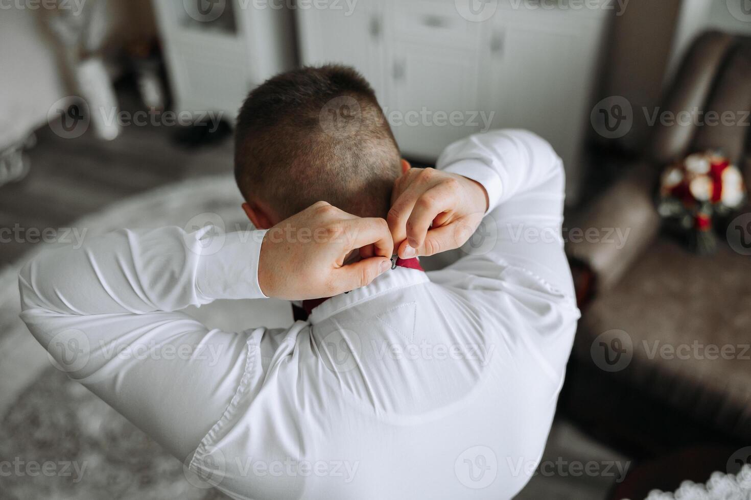a young man in a white shirt stands by the window in the room and puts on a tie. The groom gets dressed in the morning and prepares for the wedding. photo
