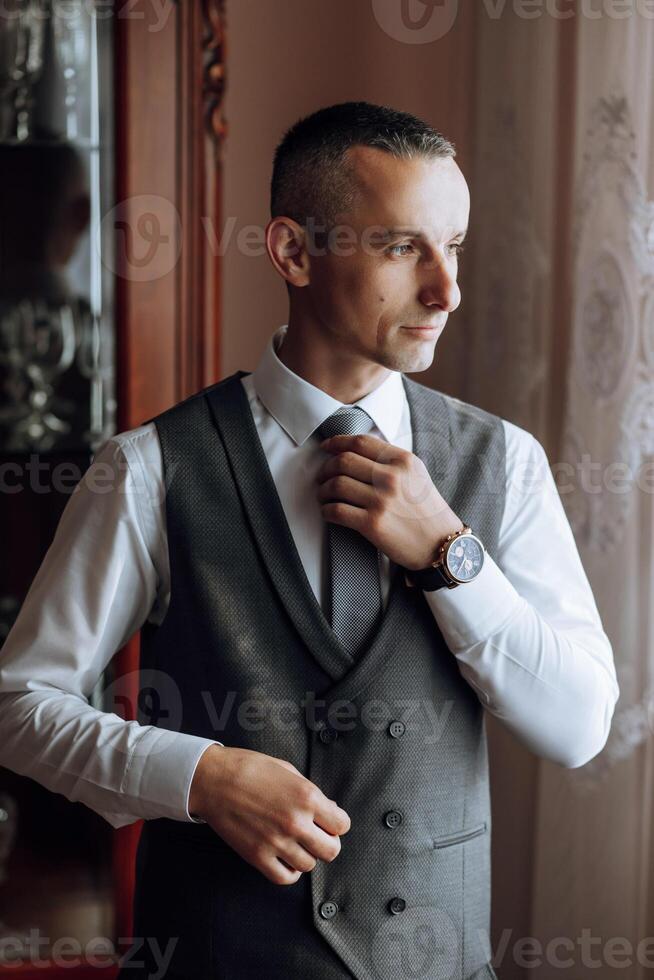 a young man in a white shirt stands by the window in the room and puts on a tie. The groom gets dressed in the morning and prepares for the wedding. photo