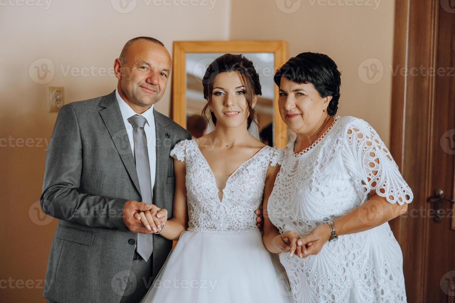 Happy parents hold the hands of the bride who is getting married and support her, wearing a wedding dress and a long veil. photo