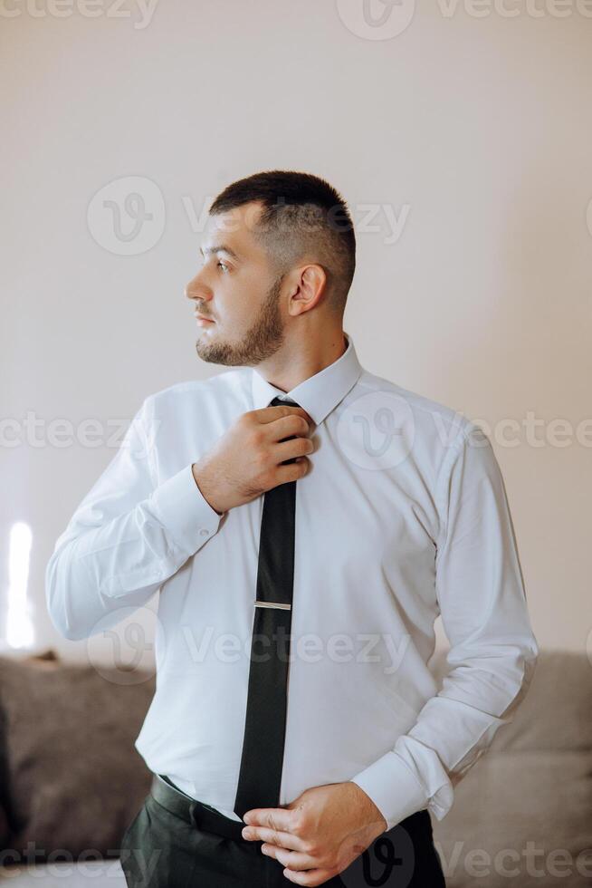 a young man in a white shirt stands by the window in the room and puts on a tie. The groom gets dressed in the morning and prepares for the wedding. photo