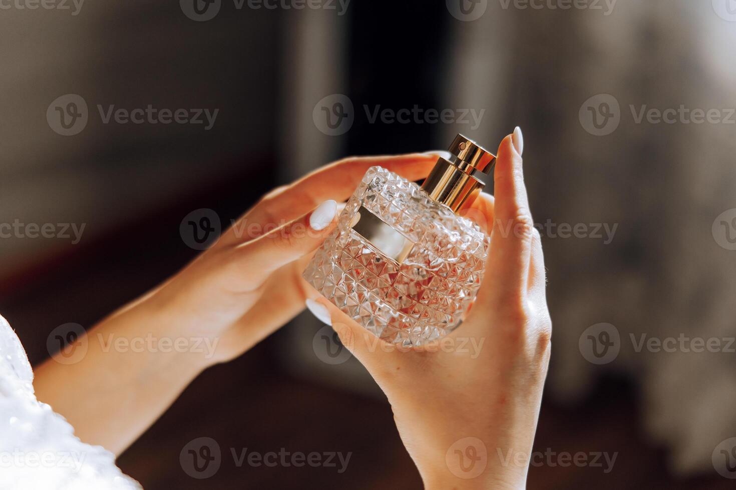 Glass bottle of perfume in gentle female hands. The bride sprays perfume on her body. Close-up photo. Manicure. Morning of the bride. photo