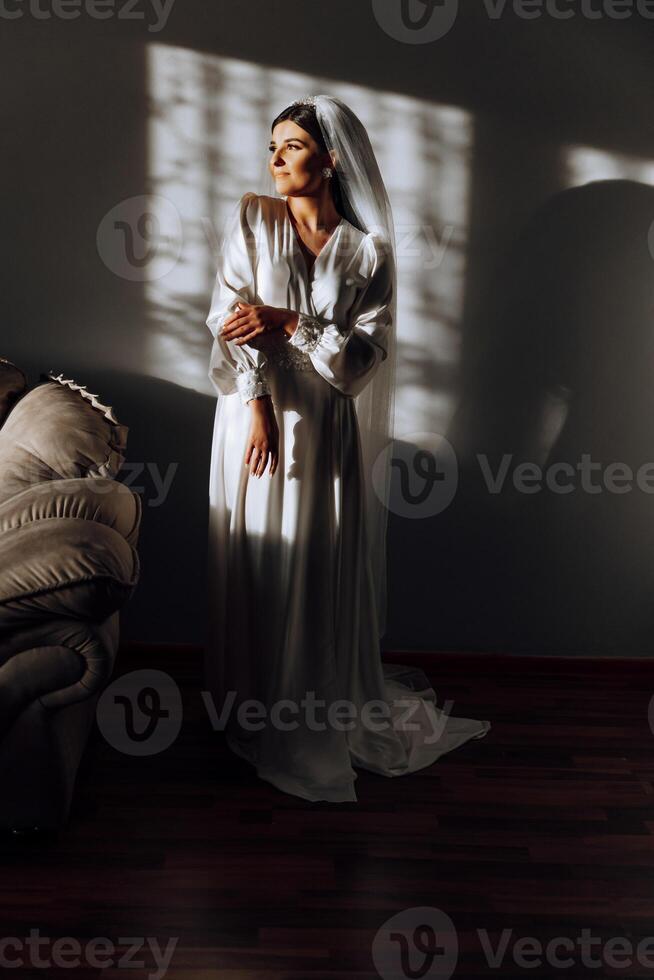 portrait of an incredibly beautiful girl bride in a white robe in the bedroom in beautiful light and shadow, the bride poses in the morning before the wedding ceremony. photo