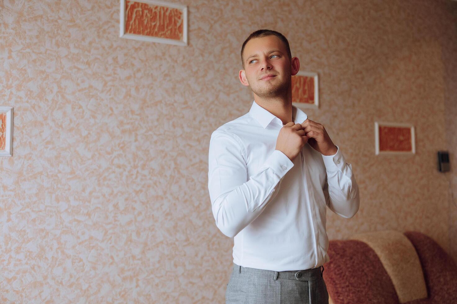 un hombre en un blanco camisa soportes por el ventana en el habitación y sujeta el botones en su collar y mangas reloj en mano. elegante negocio retrato de un hombre, de cerca foto. el novio es preparando. foto