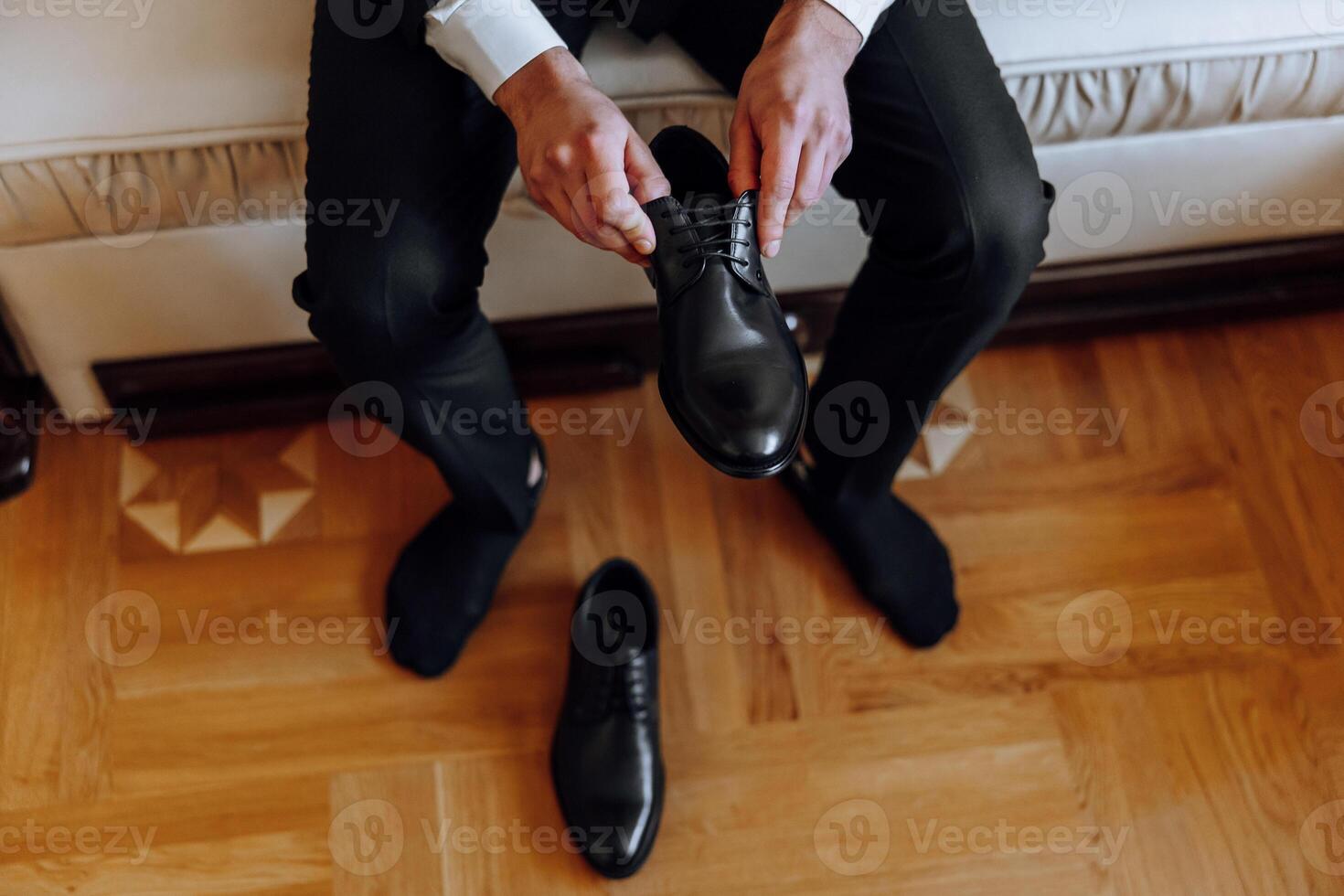 A man is putting on his shoes while sitting on a sofa in a hotel room. A young entrepreneur is preparing to go on a business trip. Businessman preparing for travel in home bedroom early in the morning photo