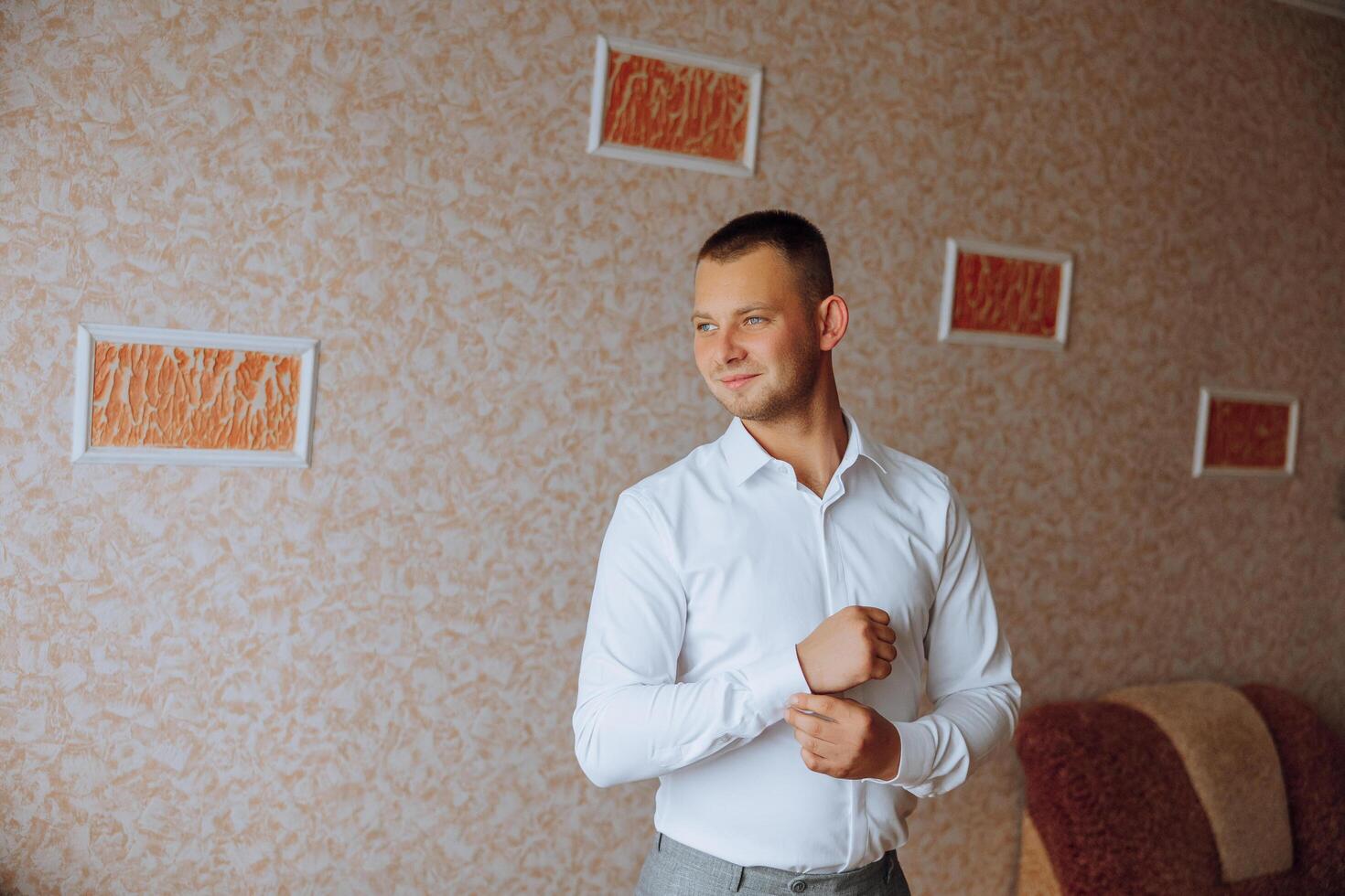 un hombre en un blanco camisa soportes por el ventana en el habitación y sujeta el botones en su collar y mangas reloj en mano. elegante negocio retrato de un hombre, de cerca foto. el novio es preparando. foto