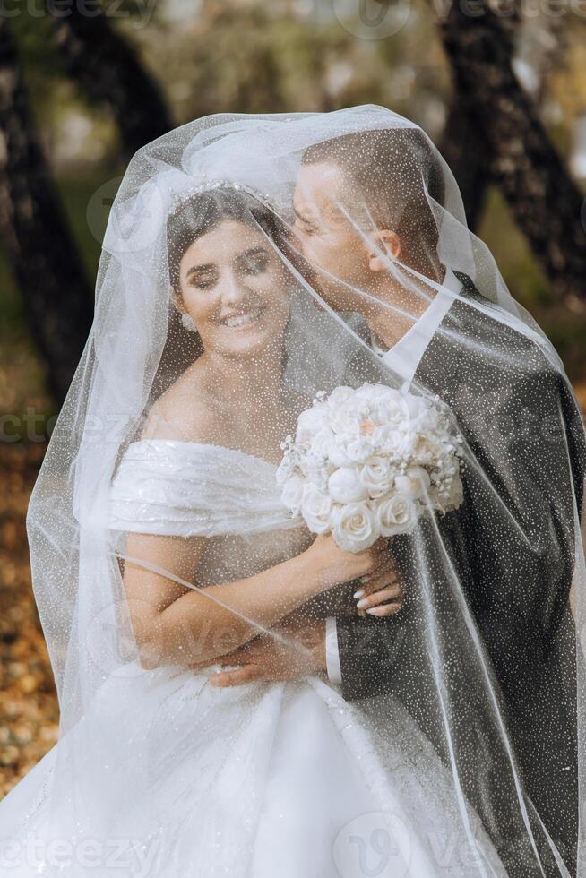 A young bride and groom tenderly embrace in the rays of the autumn sun. Tender and beautiful young girl bride. A man kisses his beloved. Against the background of a beautiful garden photo