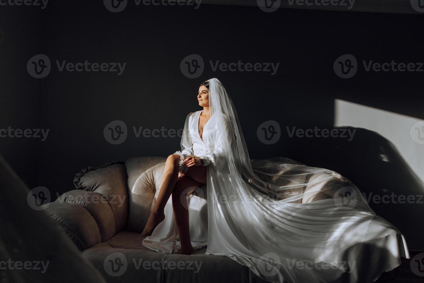 an incredibly beautiful bride in the morning in a white robe and a long veil sits on the bed in her bedroom. The bride poses in the morning before the wedding ceremony. photo