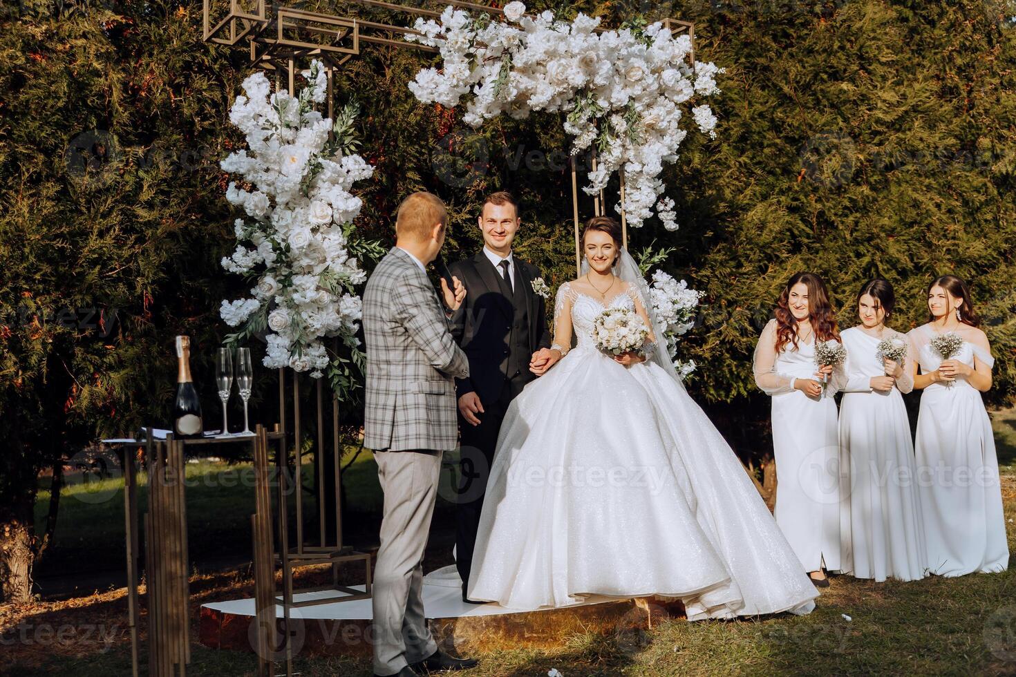 A master of ceremonies gives a speech while a bride and groom hold hands near a white floral arch photo