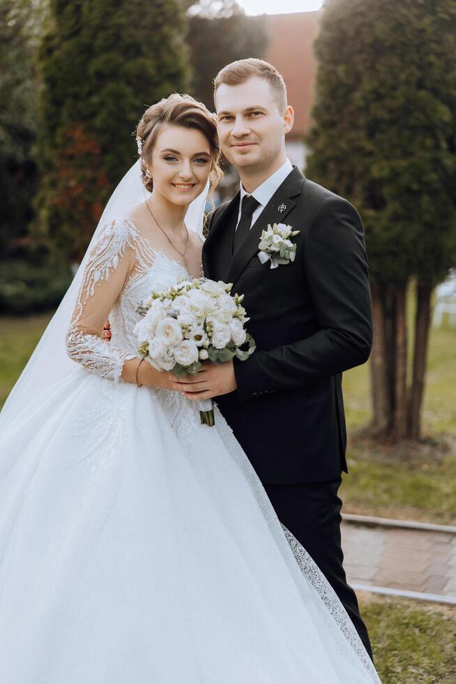 Stylish, young groom and beautiful bride in a long white dress and a long veil with a bouquet in their hands, hugging in the park in the autumn nature. Wedding portrait of newlyweds. photo