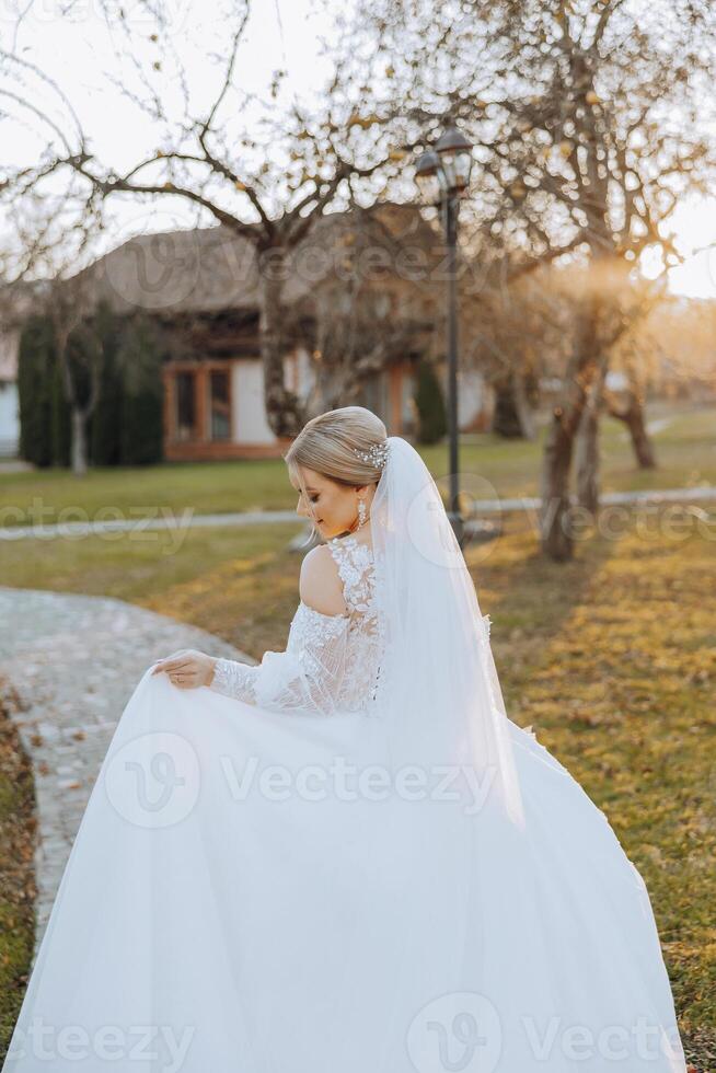 A blonde bride in a white dress with a long train holds the dress and walks along the stone path. Autumn. Wedding photo session in nature. Beautiful hair and makeup. celebration