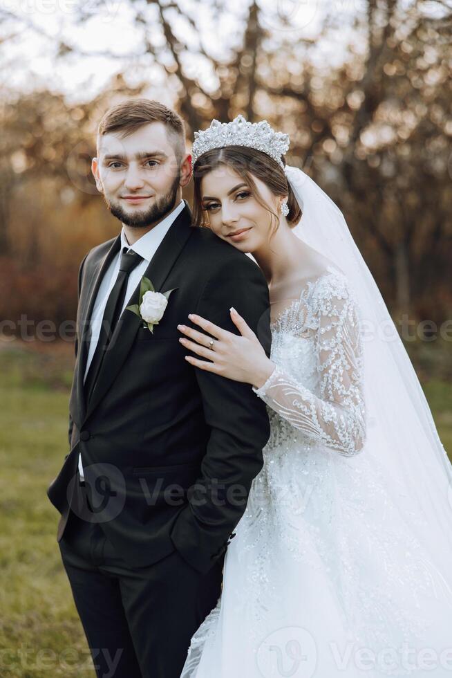 Stylish, young groom and beautiful bride in a long white dress and a long veil with a bouquet in their hands, hugging in the park in the autumn nature. Wedding portrait of newlyweds. photo