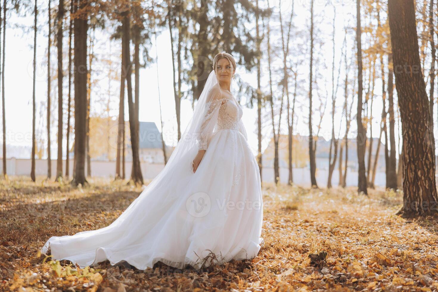 A blonde bride in a white dress with a long train holds the dress and walks down the path covered with autumn leaves. Wedding photo session in nature. Beautiful hair and makeup. Celebration