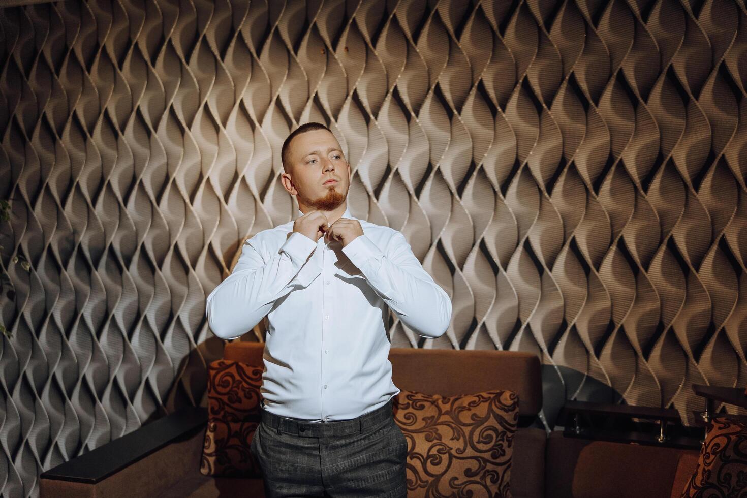 un hombre en un blanco camisa soportes por el ventana en el habitación y sujeta el botones en su collar y mangas reloj en mano. elegante negocio retrato de un hombre, de cerca foto. el novio es preparando. foto