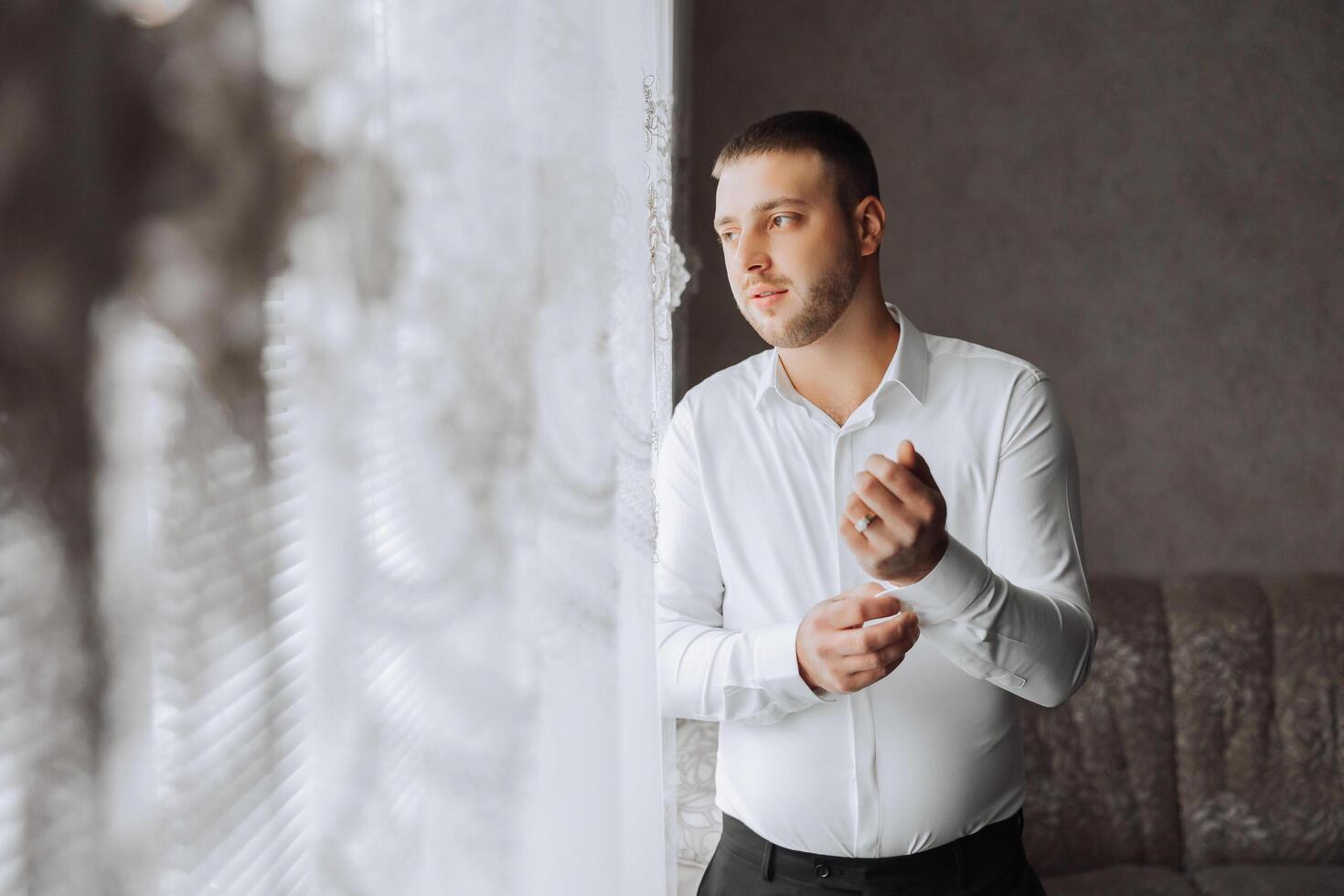 A man in a white shirt stands by the window in the room and fastens the buttons on his collar and sleeves. Watch on hand. Stylish business portrait of a man, close-up photo. The groom is preparing. photo