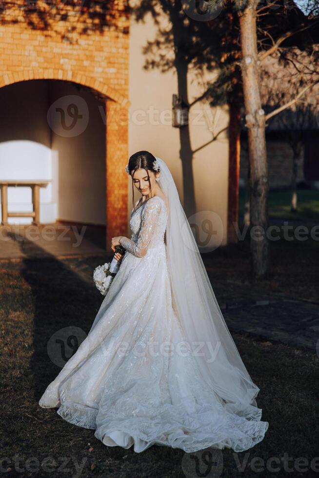 A brunette bride in a white dress with a long train holds the dress and walks down the stone path. Autumn. Wedding photo session in nature. Beautiful hair and makeup. celebration