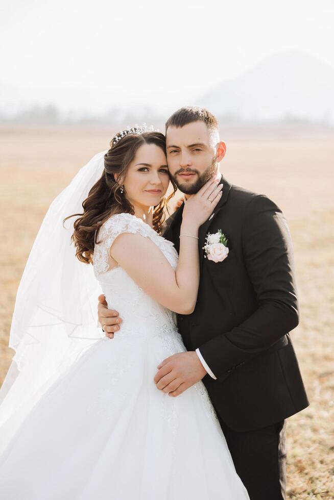 elegante, joven novio y hermosa novia en un largo blanco vestir y un largo velo con un ramo de flores en su manos, abrazando en el parque en el otoño naturaleza. Boda retrato de recién casados. foto