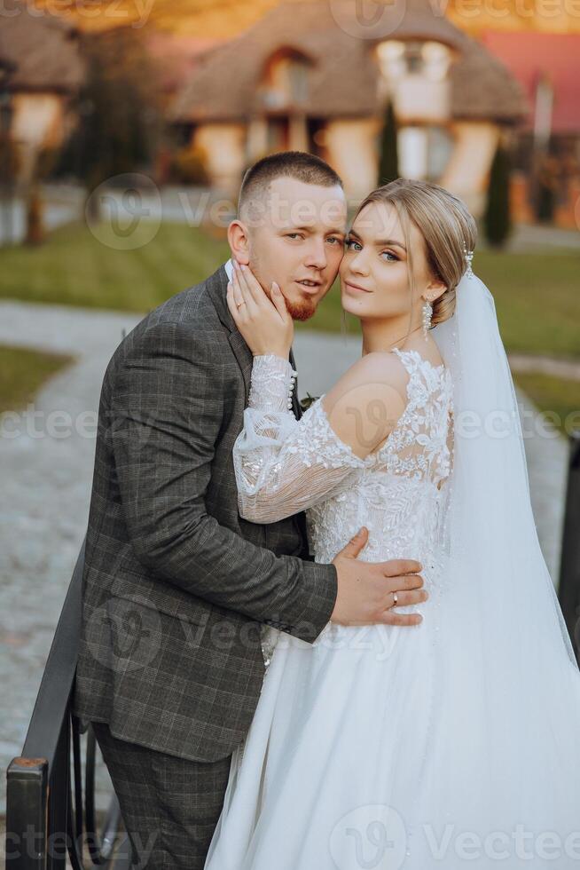 Stylish, young groom and beautiful bride in a long white dress and a long veil with a bouquet in their hands, hugging in the park in the autumn nature. Wedding portrait of newlyweds. photo
